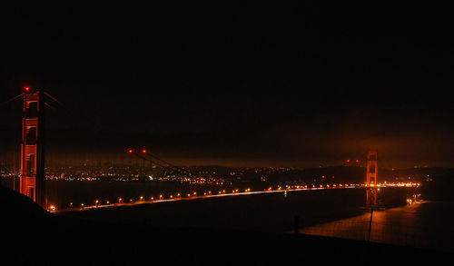 Illuminated bridge over river by buildings against sky at night