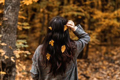 Back view of brunette woman with fall yellow leaves in long hair on autumn nature background