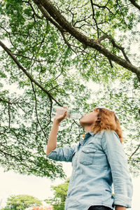 Woman drinking water from bottle while standing against trees