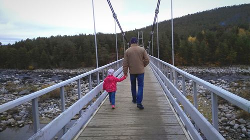 Rear view of father holding hands of daughter while walking on footbridge during winter