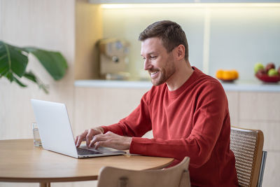 Positive cheerful man enjoying work from home as freelancer using laptop in cozy modern apartment