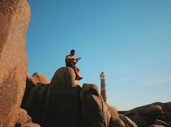 Low angle view of mid adult man playing guitar while sitting on rock formation against blue sky