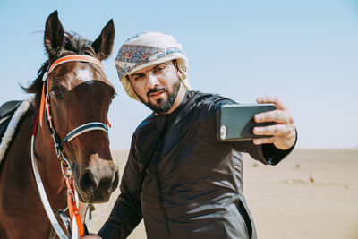 Young man taking selfie with mobile phone riding horse at desert