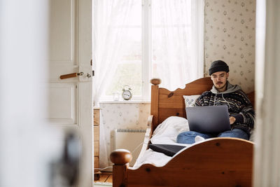 Young man using laptop while sitting on bed seen through doorway at home