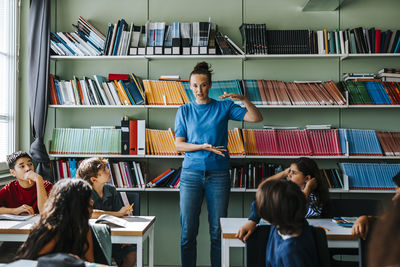 Female teacher gesturing and explaining group of elementary students while standing in front of bookshelf at library