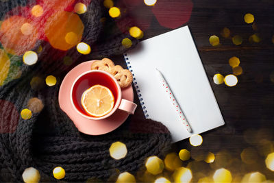 Clear copybook, cup of pomegranate tea, pen on black wooden background with christmas lights