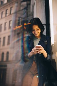 Smiling young woman texting through phone while standing by glass window