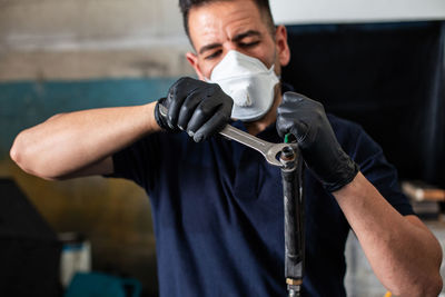 Male mechanic in respirator and latex gloves screwing nut on nozzle with spanner while working in professional workshop