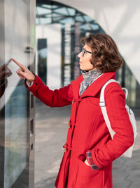 Curly woman in red coat is scrolling screen on outdoor electronic navigation device. 