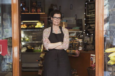 Portrait of confident owner standing with arms crossed at store seen from entrance