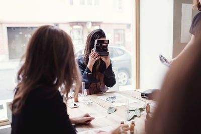 Woman photographing while sitting by colleague at table against window in workshop