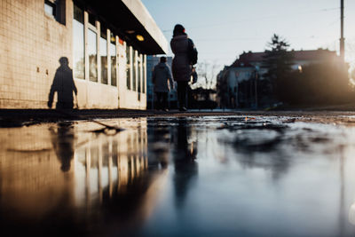 Rear view of people walking on puddle on footpath in city against sky