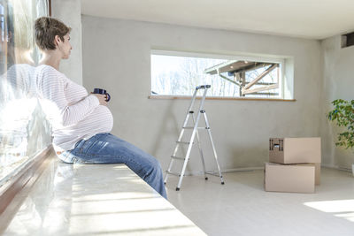 Pregnant woman holding cup while sitting by window at home