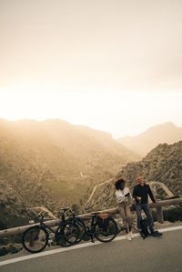 Man and woman with bicycles standing roadside at sunset on vacation