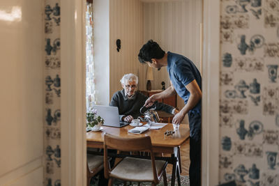 Side view of male healthcare worker serving tea to senior man at home