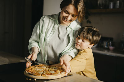 Son embracing mother holding pizza and cutter at home