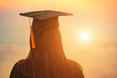 Rear view of woman wearing mortarboard while standing against orange sky
