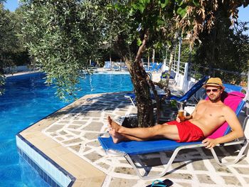 Shirtless young man holding drink relaxing on deck chair at poolside