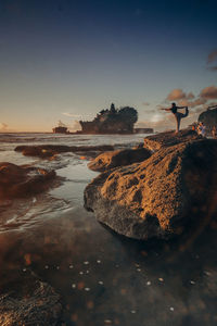 Man on rock by sea against sky during sunset