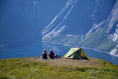 People sitting by tent on mountain peak against lake