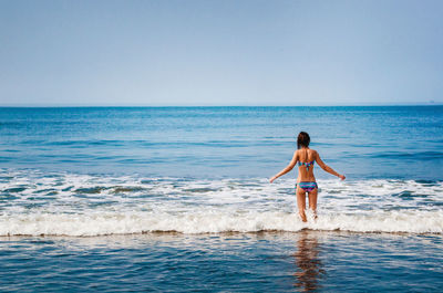 Rear view of woman with arms outstretched wearing bikini standing in sea against clear sky