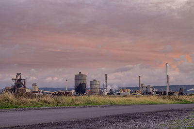 Road by factory against sky during sunset