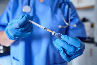 Crop anonymous medic in uniform and latex gloves withdrawing medication from vial into syringe while standing in kitchen on blurred background