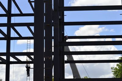 Low angle view of worker climbing ladder on metallic structure against sky