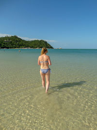 Rear view of mid adult woman in bikini walking at beach against blue sky