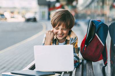 A cheerful boy is lying on a wooden bench and working on a laptop, next to a backpack. 