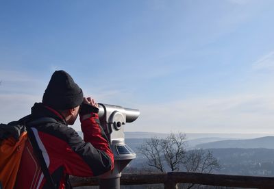 Man looking through coin-operated binoculars against sky during winter