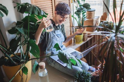 Happy young girl with laptop among plants on balcony, green environment in room, home gardening