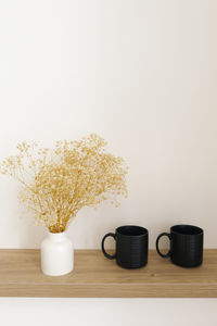 Dried flowers in a white ceramic vase and two black mugs on a wooden countertop in the decor