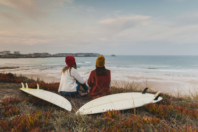 Female friends sitting with surfboards at beach during sunset