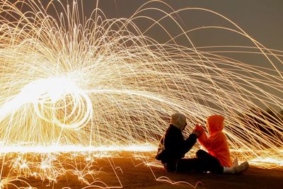 People sitting by illuminated wire wool at night against sky