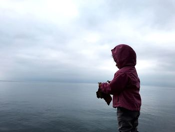 Girl wearing pink hooded shirt standing at sea against cloudy sky