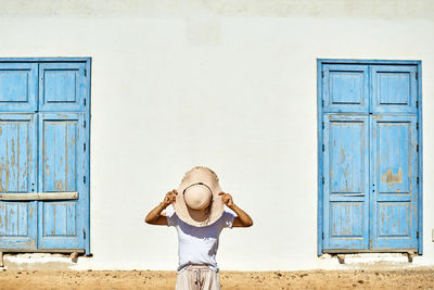 Unrecognizable female in summer clothes covering face with hat while standing outside rural white house with blue window shutters in sunny country