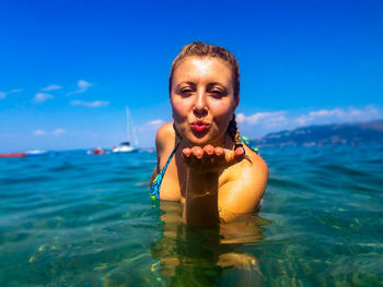 Portrait of smiling woman blowing kiss in sea against sky during sunny day