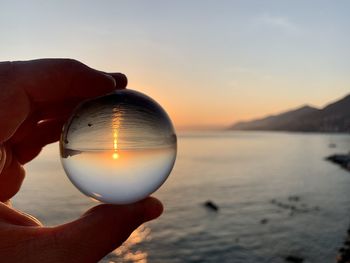 Close-up of hand holding crystal ball against sky during sunset