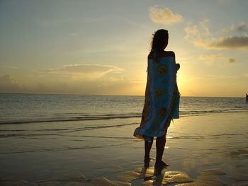 Rear view of woman wrapped in sarong while standing at sea shore during sunset