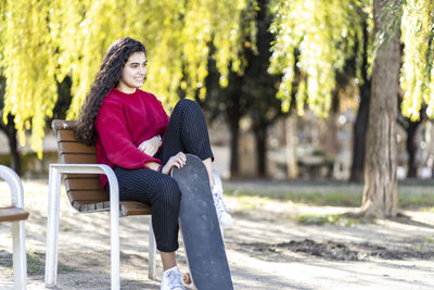 Smiling female teenager sitting on chair with skateboard in park
