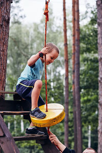 Father helping son while sitting on zip line against trees