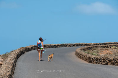 Rear view of mid adult woman with dog walking on road against blue sky