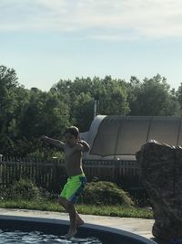 Full length of shirtless boy standing by trees against sky