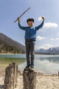 Full length portrait of boy holding stick while standing on wooden post against lake and sky