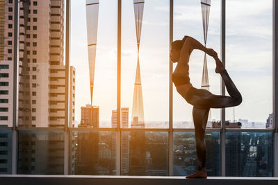 Side view of young woman exercising by window against cityscape
