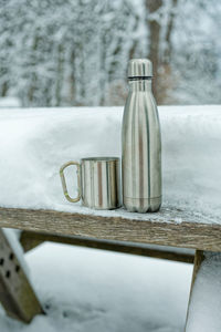 On a table covered with snow in a park stands a silver thermos flask with a metal drinking cup.