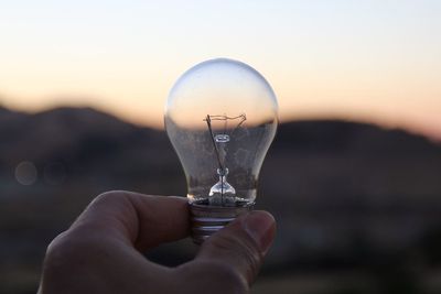 Close-up of hand holding light bulb against sky