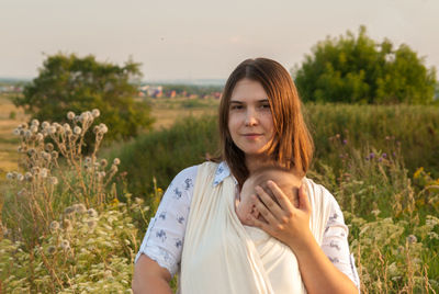 Portrait of woman with baby carrying in fabric while standing on field against sky
