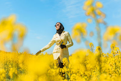 A young stylish woman poses among a blooming yellow field of rapeseed under a bright spring sky